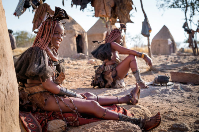 Namibia beste Reisezeit für Festivals und Kultur zum Beispiel Himba-Frauen sitzen vor ihren Hütten und kleiden sich im traditionellen Stil in einem Himba-Dorf in der Nähe von Kamanjab - credit: R.M. Nunes - Stock-Fotografie-ID:1429339469