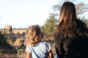 Young girl sits questions older sister about the elephant they are watching in the distance